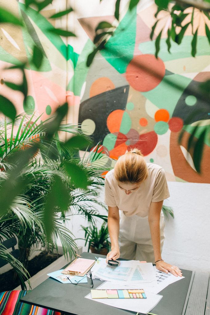 A focused woman reviews documents in a lush, colorful workspace adorned with abstract art.