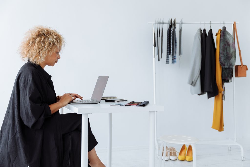 Businesswoman working on a laptop at a white desk with a clothing rack in a bright, minimalist office.
