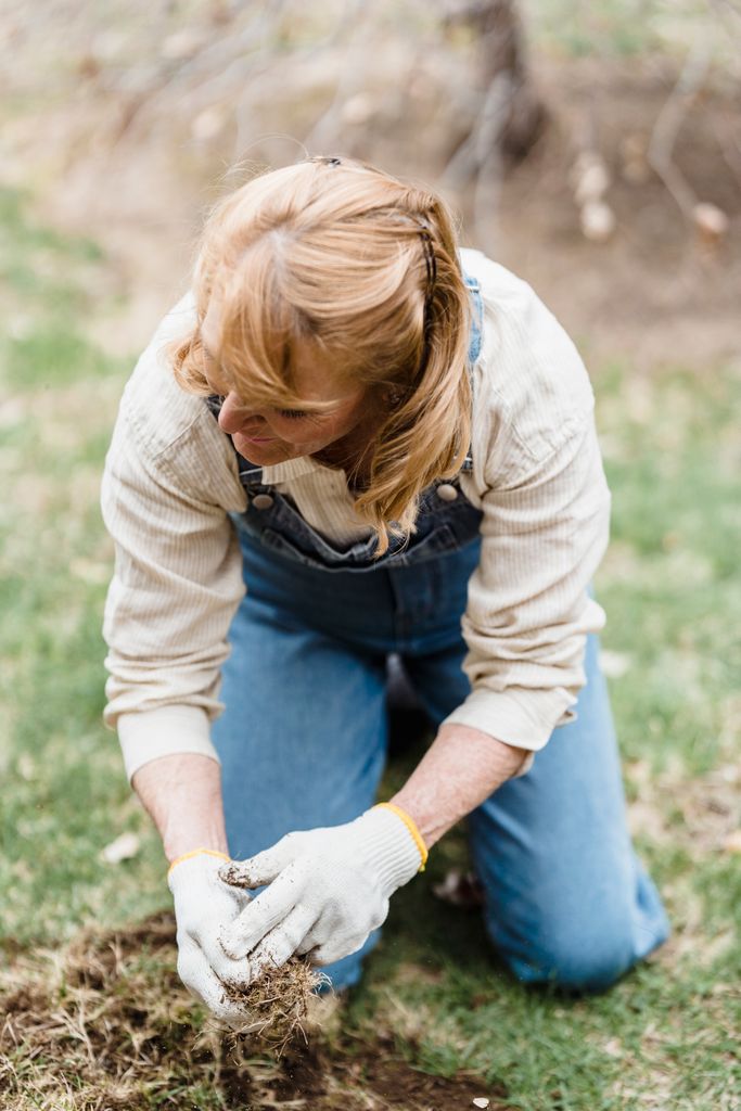 Full body of unrecognizable female gardener in gloves sitting on grassy ground while working in garden